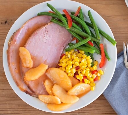 Slices of ham, corn, fresh green beans and apples on a dinner plate.