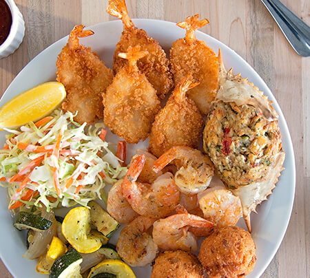 A plate of seafood including butterfly shrimp, crab cakes, gulf shrimp and hush puppies