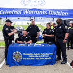 Several police officers in a tent with the sign "Criminal Warrants Division" on the top. One of them is laughing.